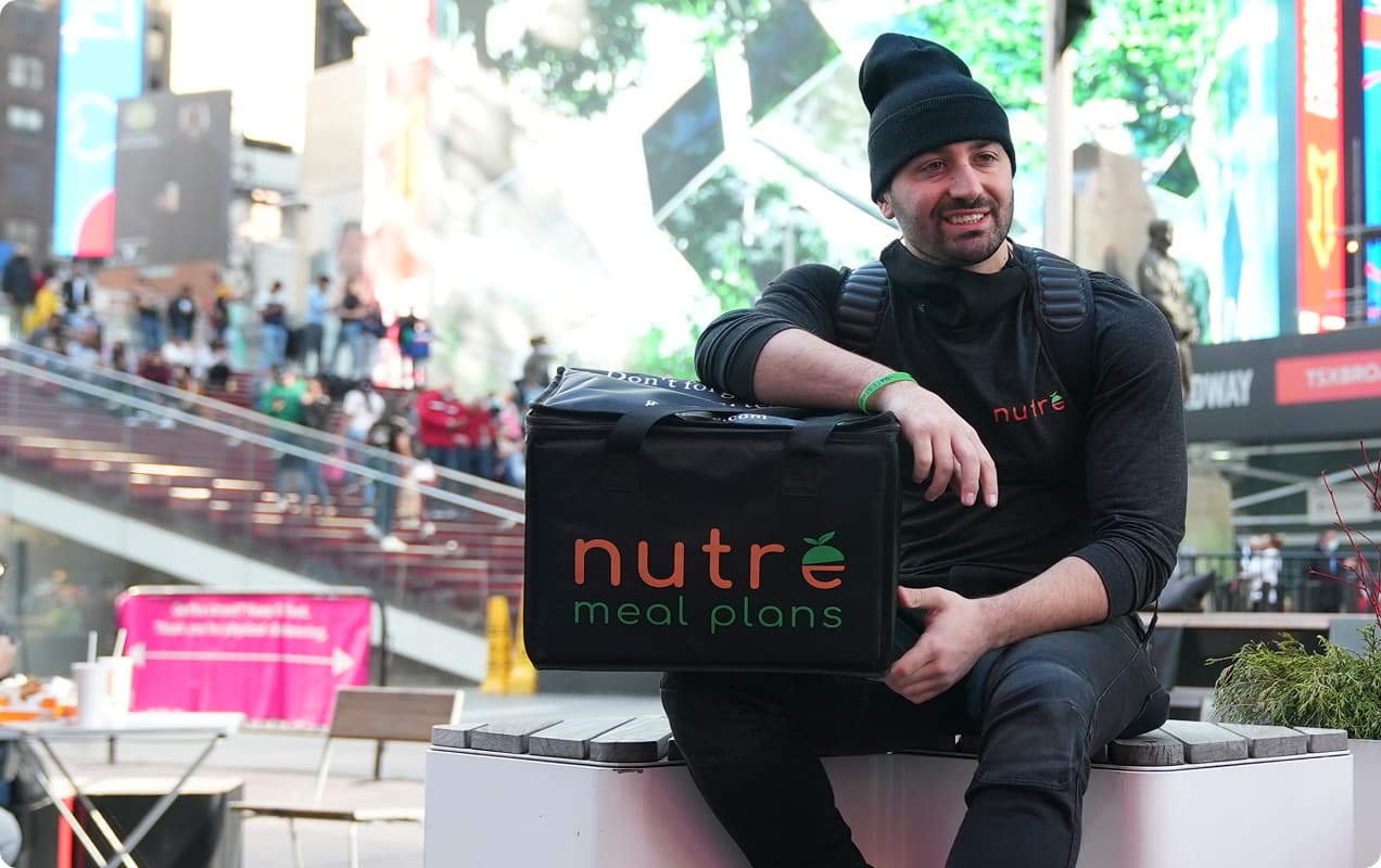 Man in black beanie smiling, holding Nutre meal delivery bag in urban setting.